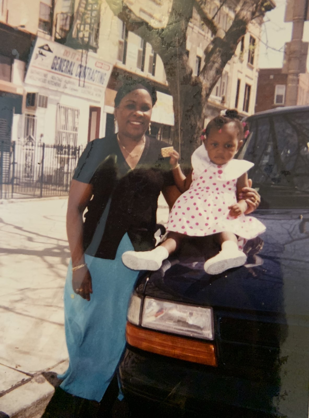 Lilianne smiling on a Brooklyn sidewalk beside the founder as a toddler in a pink polka-dot dress, sitting on the hood of a car